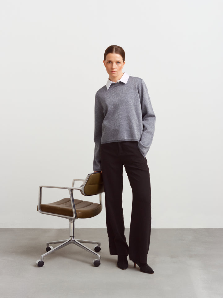 Montreal sweater in gun metal glow, seated by a chair with hands in trouser pockets